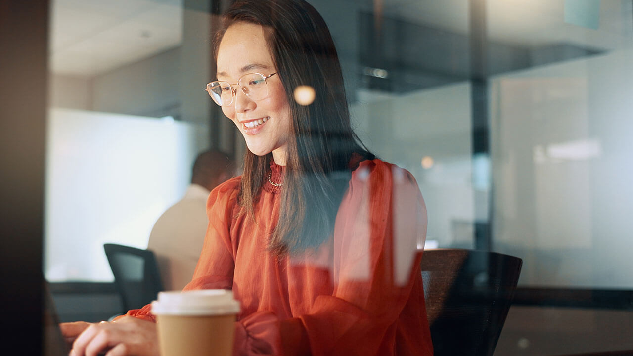 A woman in a red blouse smiles while working on a laptop in a modern office setting.